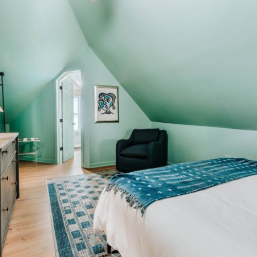 Settle into this inviting bedroom with crisp linens, a wall-mounted TV, comfortable lounge chair, and convenient dresser storage. Unique sloped ceilings and warm lighting create a relaxing retreat for movie nights and restful sleep.