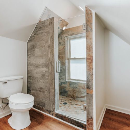 Step into a bright, modern bathroom featuring a beautifully tiled walk-in shower with a glass enclosure and warm wood flooring. The unique sloped ceilings add character, creating a refreshing space to start your day or unwind at night.