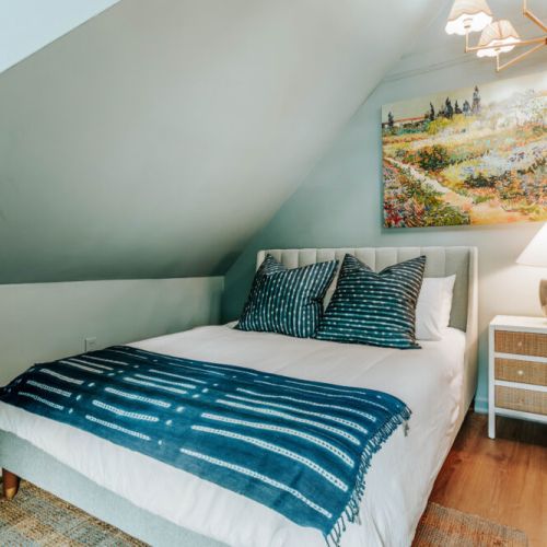 Unwind in this serene bedroom beneath charming sloped ceilings, with crisp linens, rich blue accents, warm bedside lighting, and colorful artwork. It’s a peaceful retreat designed for restful nights and slow, easy mornings.